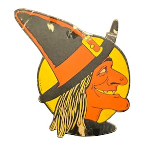 May include: Vintage Halloween decoration featuring a smiling witch's face. The witch has orange skin, a large nose, and yellow hair. She wears a black and orange witch hat against a yellow background. The decoration is made of cardboard.
