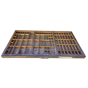 May include: A vintage wooden printer's tray with numerous small compartments of varying sizes. The tray is rectangular, with a dark, weathered finish. It is designed to hold individual pieces of type for letterpress printing.