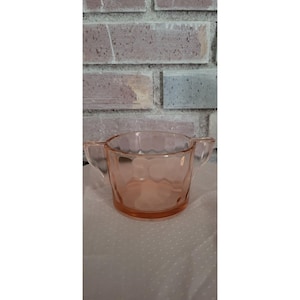 Puede incluir: Un azucarero de cristal rosa con dos asas. El cuenco tiene un patrón texturizado y está colocado sobre un fondo de pared de ladrillo. El cristal es de un rosa suave y translúcido.