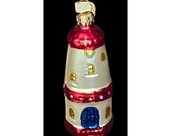 Könnte beinhalten: Ein weihnachtlicher Leuchtturm-Ornament. Das Ornament hat einen weißen Körper mit roten Akzenten an der Ober- und Unterseite. Es hat kleine, rechteckige Fenster und eine blaue Tür. Das Ornament ist etwa 10 cm hoch.