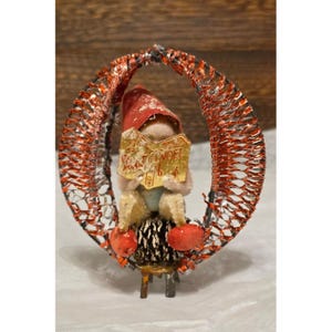 May include: A vintage Christmas ornament featuring a small gnome reading a book with the word "NOEL" on it. The gnome is seated on a black and gold base, surrounded by a red and silver oval decoration. Two red spheres are on the base.