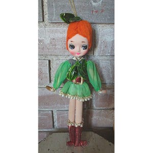 Puede incluir: Muñeca vintage con cabello de hilo naranja, vestida con un atuendo verde con un cuello y cinturón en forma de hoja a juego. La muñeca tiene mejillas rosadas, ojos grandes y botas rojas. Se adjunta una cuerda dorada para colgar.