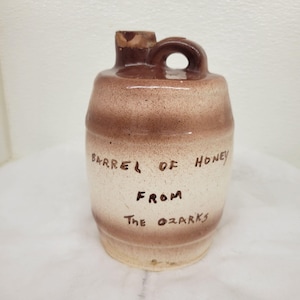 May include: A brown and white ceramic honey jar with the text "Barrel of Honey From The Ozarks" written on it.