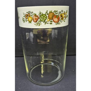 May include: A clear glass jar with a white band featuring a colourful fruit and vegetable design. The jar is cylindrical and appears to be empty. The design includes images of tomatoes, lemons, and other produce.