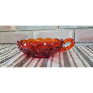 May include: A vintage, ruby red glass dish with a handle. The dish has a scalloped rim and is decorated with starburst patterns. The handle is curved and the glass has a glossy finish. The dish is likely used for serving or display.