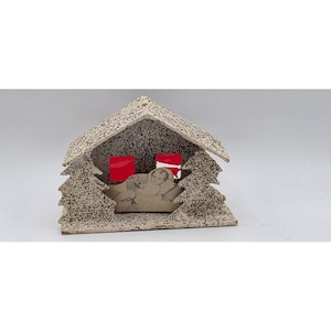 Puede incluir: Un belén navideño vintage. La estructura es una pequeña casa con un tejado texturizado y moteado y árboles de pino recortados a ambos lados. En el interior, una imagen impresa representa al niño Jesús. Se ven dos rectángulos rojos.