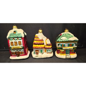 May include: Three glass Christmas ornaments shaped like miniature buildings. The ornaments feature detailed facades with snow-covered roofs and accents. One reads "WAREHOUSE" and another reads "S. COUGHLEY".