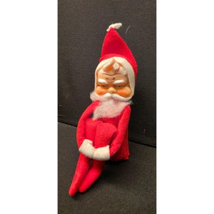 May include: A plush Santa Claus doll featuring a bright red pointed hat and matching red suit with white trim. The doll has a moulded face with painted eyes, a white synthetic beard, and white mittens, depicted in a seated position with knees pulled up.