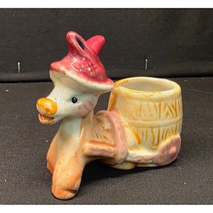 Może przedstawiać: Ceramiczna figurka osła z doniczką w kształcie beczki. Osiołek pomalowany w odcieniach bieli, pomarańczu i różu, w czerwonym kapeluszu. Doniczka w kształcie beczki jest jasnożółta z brązowymi akcentami.