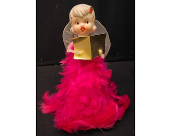 May include: A decorative figurine of a choir angel with blonde hair and a red hair accessory. The angel has a translucent wing structure and is holding a gold-colored book. The body is covered in vibrant, bright pink feathers in a skirt shape.
