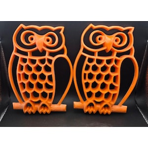 May include: Two orange owl-shaped decorations. Each owl is perched on a branch and has a hollow, honeycomb-like body. The owls have large, round eyes and a pointed beak. The background is black.