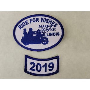Op de afbeelding: Twee witte geborduurde patches met blauwe stiksels. De grotere patch is ovaalvormig en zegt "Ride for Wishes Make-A-Wish Illinois". De kleinere patch is een gebogen rechthoek met het jaar "2019" in blauw.
