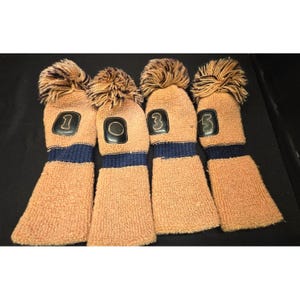 Puede incluir: Juego de cuatro fundas para palos de golf. Las fundas son de color marrón claro con una franja azul oscuro en la parte inferior. Cada funda tiene un número diferente: 1, 0, 3 y 3. La parte superior de cada funda tiene un pompón marrón.