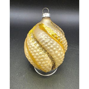 May include: A pear-shaped glass Christmas ornament with a textured, dimpled surface in a pale cream color. It features diagonal bands of bright yellow paint and thin lines of dark gold glitter. A silver metal cap with a hanging loop is at the top.