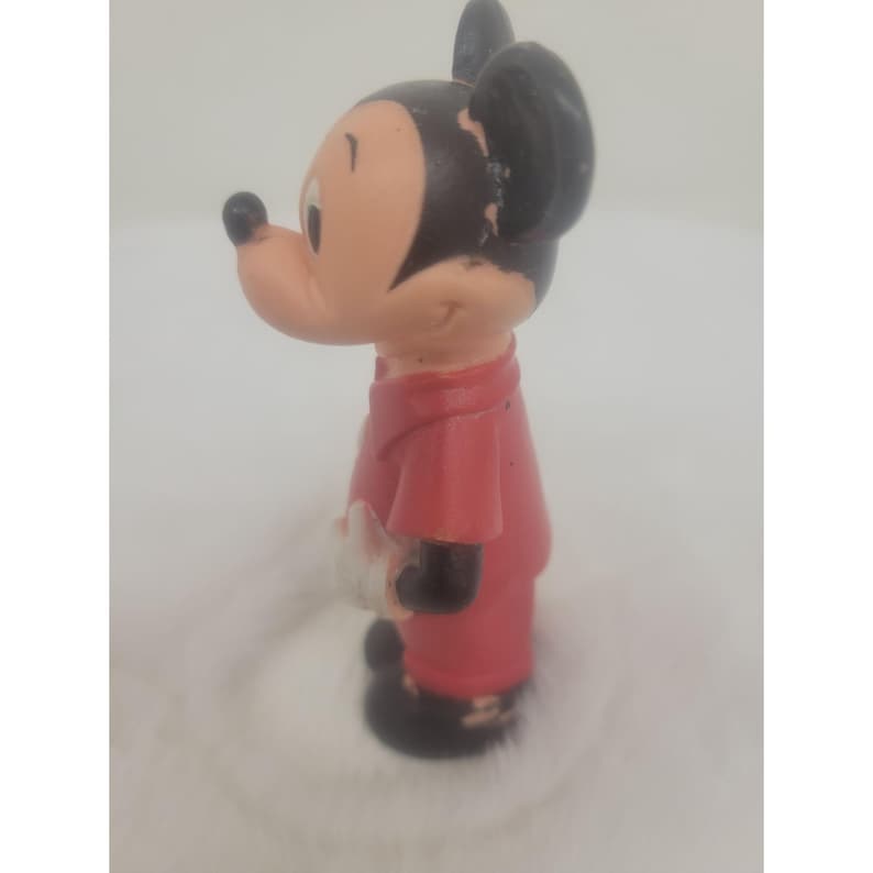 May include: A red and black plastic figurine of Mickey Mouse wearing a red shirt and black shoes. He is holding a white object in his right hand.