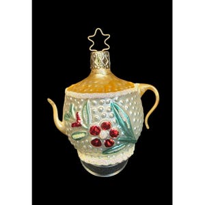May include: A glass teapot-shaped Christmas ornament featuring a textured, dimpled surface in a pale silver finish. The design includes painted red flowers with green leaves, a gold-toned top section, and a star-shaped wire hanger at the peak.