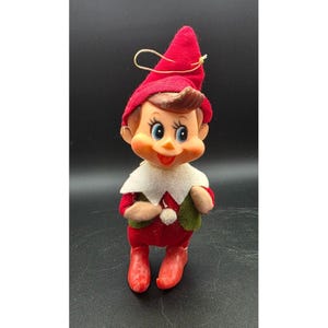 May include: A vintage elf figurine featuring a red felt hat with a gold hanging loop, a white felt collar, a green felt vest, and red pointed boots. The elf has a moulded plastic face with large blue eyes, brown hair, and a smiling expression.