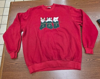 Könnte beinhalten: Ein rotes Langarm-Sweatshirt mit Rundhalsausschnitt und einer Grafik von drei Katzen, die aus drei grünen Weihnachtsstrümpfen schauen. Jeder Strumpf ist mit einem Buchstaben bestickt, die zusammen das Wort JOY in Gold ergeben.