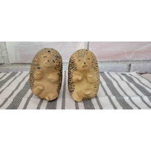 May include: A pair of ceramic hedgehog salt and pepper shakers. The hedgehogs are tan with black markings and small, smiling faces. They have small arms and legs. The shakers are on a striped cloth.