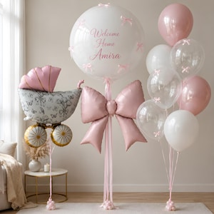 May include: A balloon arrangement featuring a large clear balloon with the text Welcome Home Amira and small pink bows, a pink foil bow, a baby carriage-shaped balloon with a floral pattern, and a cluster of pink and white balloons.