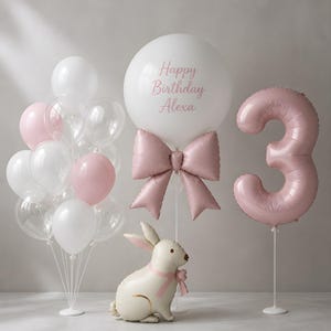 May include: A balloon arrangement featuring a large white balloon with the text Happy Birthday Alexa, a pink bow balloon, a pink number 3 balloon, a cluster of white and pink balloons, and a cream-colored rabbit-shaped foil balloon.