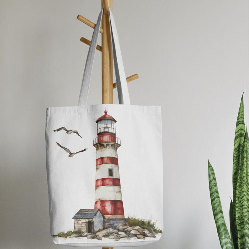 May include: White tote bag featuring a watercolor illustration of a lighthouse with red and white stripes, a small stone building, and two seagulls in flight. The lighthouse has a red top and is set on a rocky base. The bag has long, white handles.