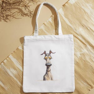May include: White tote bag featuring a whimsical illustration of a dog with large, expressive eyes and a long snout. The dog is depicted in shades of gray and tan, with a red collar. The bag has white handles.