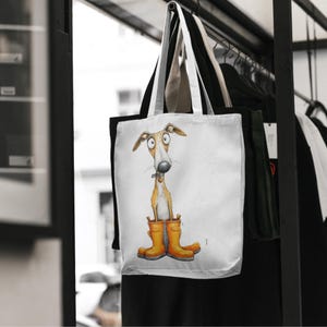 May include: White tote bag featuring a whimsical illustration of a dog wearing orange rain boots. The dog has a long snout and large eyes. The bag has a black and white handle and is hanging on a rack with other clothing.