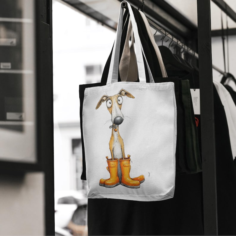 May include: White tote bag featuring a whimsical illustration of a dog wearing orange rain boots. The dog has a long snout and large eyes. The bag has a black and white handle and is hanging on a rack with other clothing.