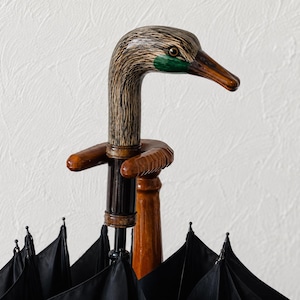 Puede incluir: Un paraguas con un mango de madera tallada en forma de cabeza de pato. La cabeza de pato presenta texturas de grano de madera marrón y beige con una mancha verde cerca del ojo y un pico marrón. La cubierta del paraguas es de tela negra.