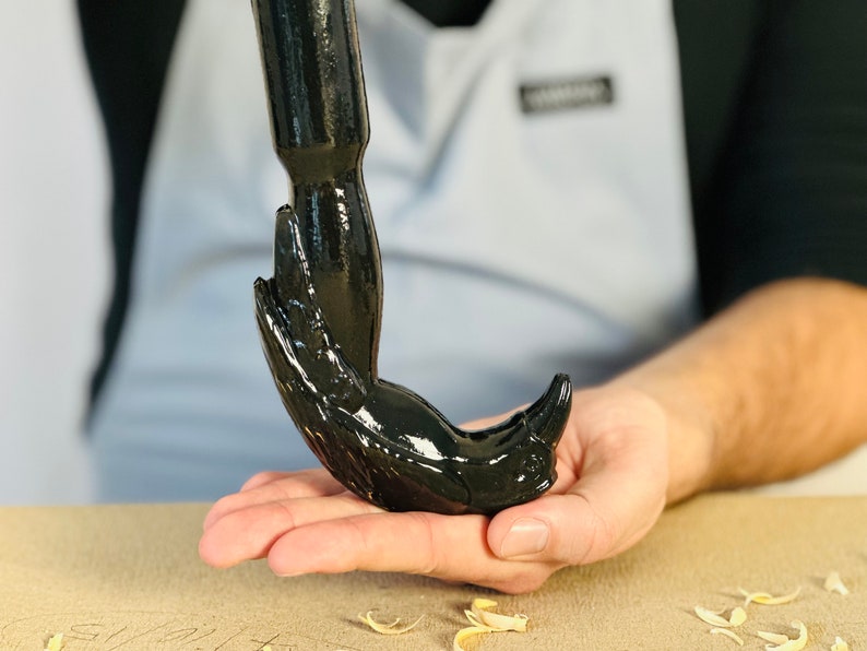 May include: A black metal hook with a bird-like design. The hook is held in a person's hand.