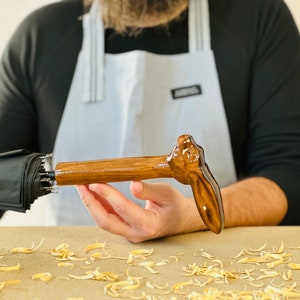 Puede incluir: Un mango de paraguas de madera tallado en forma de cabeza de conejo. El mango es de color marrón oscuro y tiene un acabado liso. El mango está siendo sostenido por la mano de una persona.