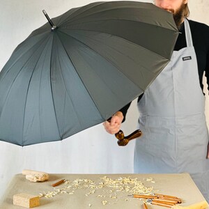 May include: A person holding a large black umbrella with a wooden handle. The umbrella is open and the person is standing in front of a white wall.
