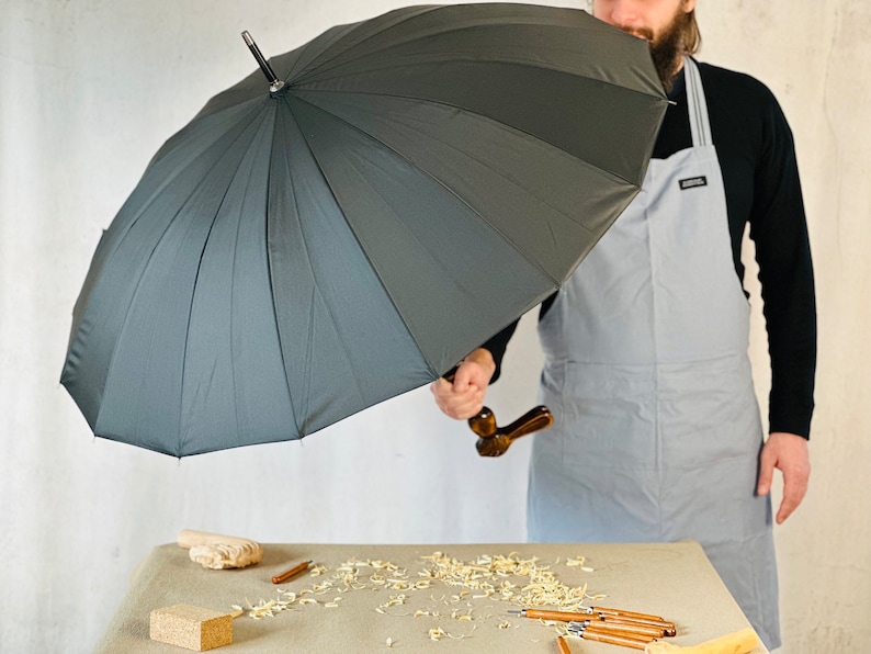 May include: A person holding a large black umbrella with a wooden handle. The umbrella is open and the person is standing in front of a white wall.