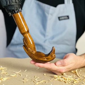May include: A wooden umbrella handle carved in the shape of a bird's head. The handle is brown and has a glossy finish. The bird's head is facing to the right and has a long beak. The handle is being held by a person's hand.