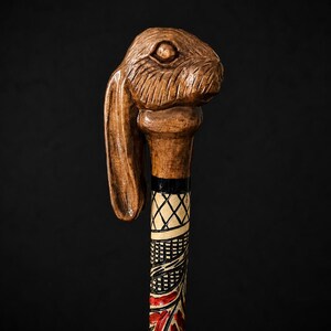 Può includere: Un bastone da passeggio con un manico in legno intagliato a forma di testa di coniglio con orecchie lunghe. L'asta sotto il manico è decorata con un motivo a griglia bianco e nero, seguito da una sezione con dettagli dipinti in rosso e nero.