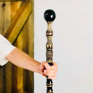 May include: A black and gold walking cane with a decorative design. The top of the cane has a black sphere, and the shaft features intricate carvings and gold accents. The cane is held by a person wearing a white shirt.