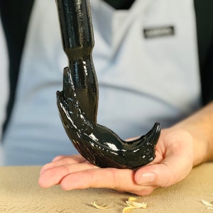 May include: A black metal hook with a unique design resembling a bird's head and beak. The hook is held in a person's hand.