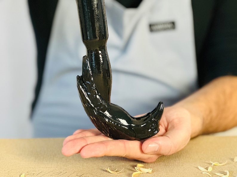 May include: A black metal hook with a unique design resembling a bird's head and beak. The hook is held in a person's hand.