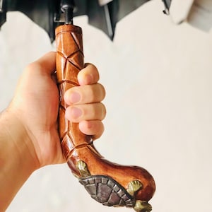 May include: A brown wooden umbrella handle with a carved geometric design and a turtle-shaped base. The handle is held by a hand, highlighting the detailed craftsmanship. The umbrella's black canopy is partially visible.