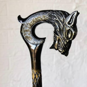 Pode incluir: Uma bengala preta e dourada com um design de cabeça de lobo. A bengala apresenta uma escultura detalhada da cabeça de um lobo no topo, com uma pega curva e um eixo inferior padronizado. O esquema de cores é principalmente preto com detalhes dourados.