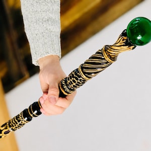 Könnte beinhalten: Ein dekorativer Stab mit einer glänzenden, kugelförmigen grünen Glaskugel an der Spitze. Der Schaft ist schwarz mit komplexen, erhabenen goldfarbenen Details, einschließlich gedrehter Spiralmuster und texturierter, schuppenartiger Segmente.