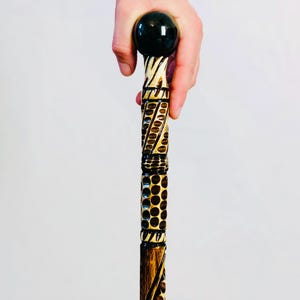 May include: A decorative walking cane with a dark, round handle. The shaft is intricately carved with geometric patterns and a dark brown finish. The cane is held by a hand against a white background.