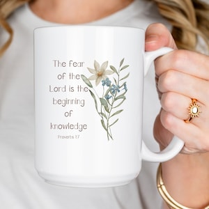 May include: White ceramic mug featuring the quote "The fear of the Lord is the beginning of knowledge" with floral illustrations. The mug has a large handle and is held by a person.