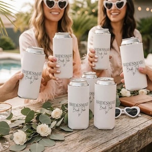 May include: White slim can coolers featuring the black printed text BRIDESMAID Bride Tribe with a small heart symbol. These cylindrical beverage insulators are designed to fit tall, slender cans and are displayed in a group setting.