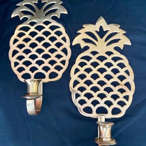 May include: Two gold-colored pineapple-shaped wall sconces. Each sconce has a detailed pineapple design with a candle holder. The sconces are made of metal and are mounted on a dark blue background.