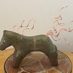 May include: A handcrafted sculpture of a horse with a green and brown body, copper wire mane and tail, and a turquoise eye. The horse is mounted on a mosaic tile base, creating a unique decorative piece.