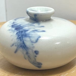 May include: A small, round, white ceramic vase with a narrow neck. The vase features a blue, abstract design that appears to be painted on the side. The vase is sitting on a wooden surface.