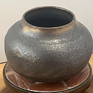 Puede incluir: Un jarrón de cerámica gris oscuro hecho a mano con una superficie texturizada y un borde dorado metálico. El jarrón redondo se asienta sobre una base decorativa multicolor. El jarrón tiene una abertura ancha y un cuerpo bulboso, adecuado para la decoración del hogar.