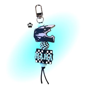 May include: A keychain featuring a black and light purple motocross helmet charm, a checkered ball, and a rectangular charm that says "MAMA" in light blue letters. The keychain has a silver clasp and light blue beads.